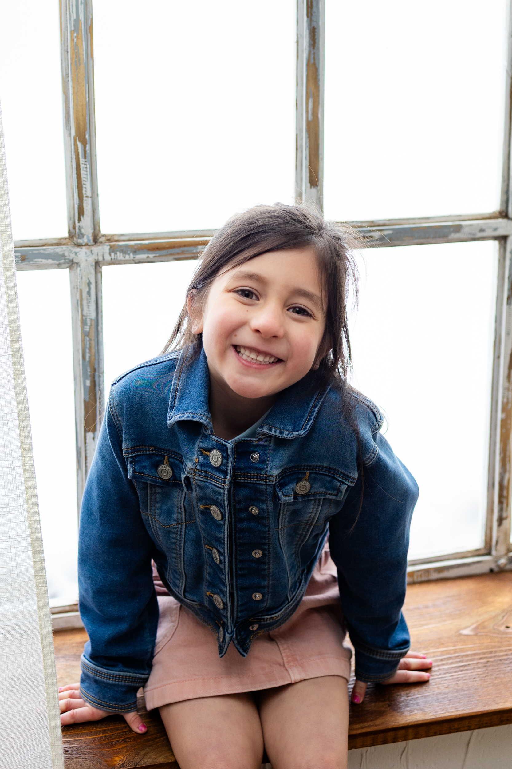 A young girl sits in a windowsill and smiles at the camera.