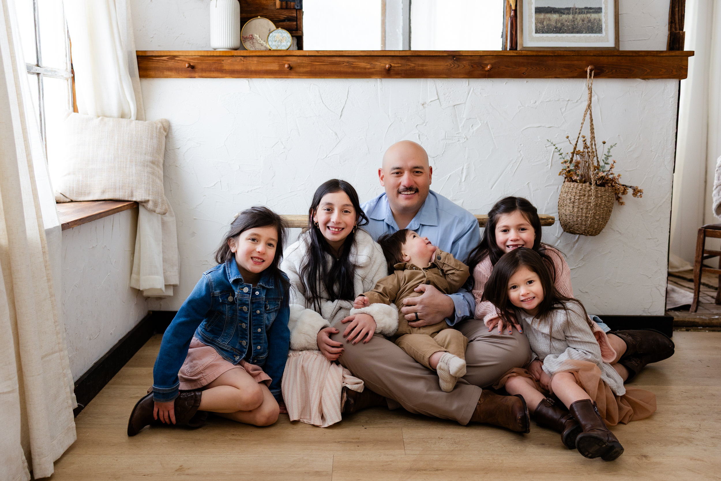 A dad sits on the floor with his five young children and they smile at the camera.