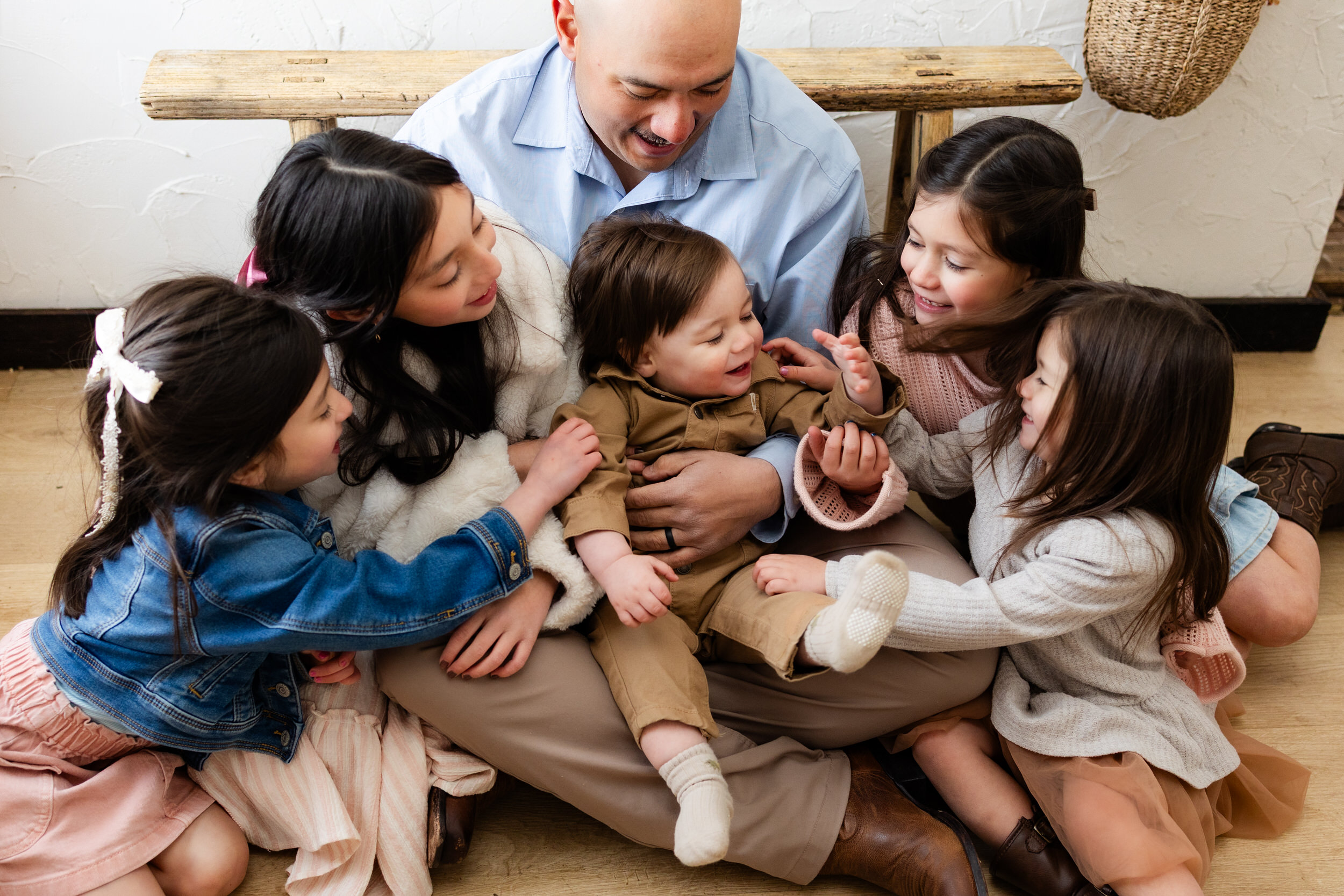 A dad sits on the floor with his five young children and they all look at the one-year-old boy sitting in his lap.