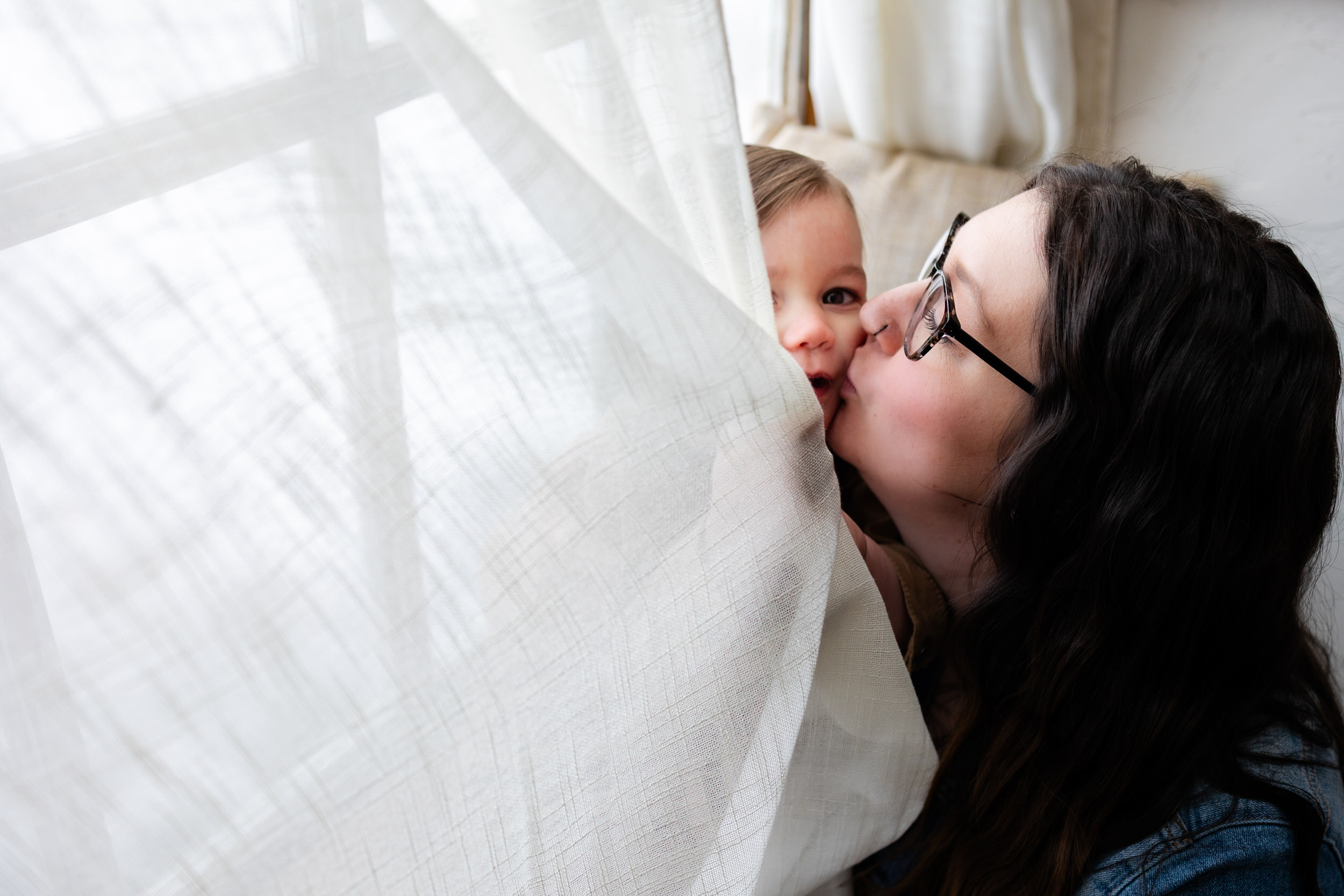 A little boy sits in a windowsill holding onto the curtain as mom kisses his cheek.