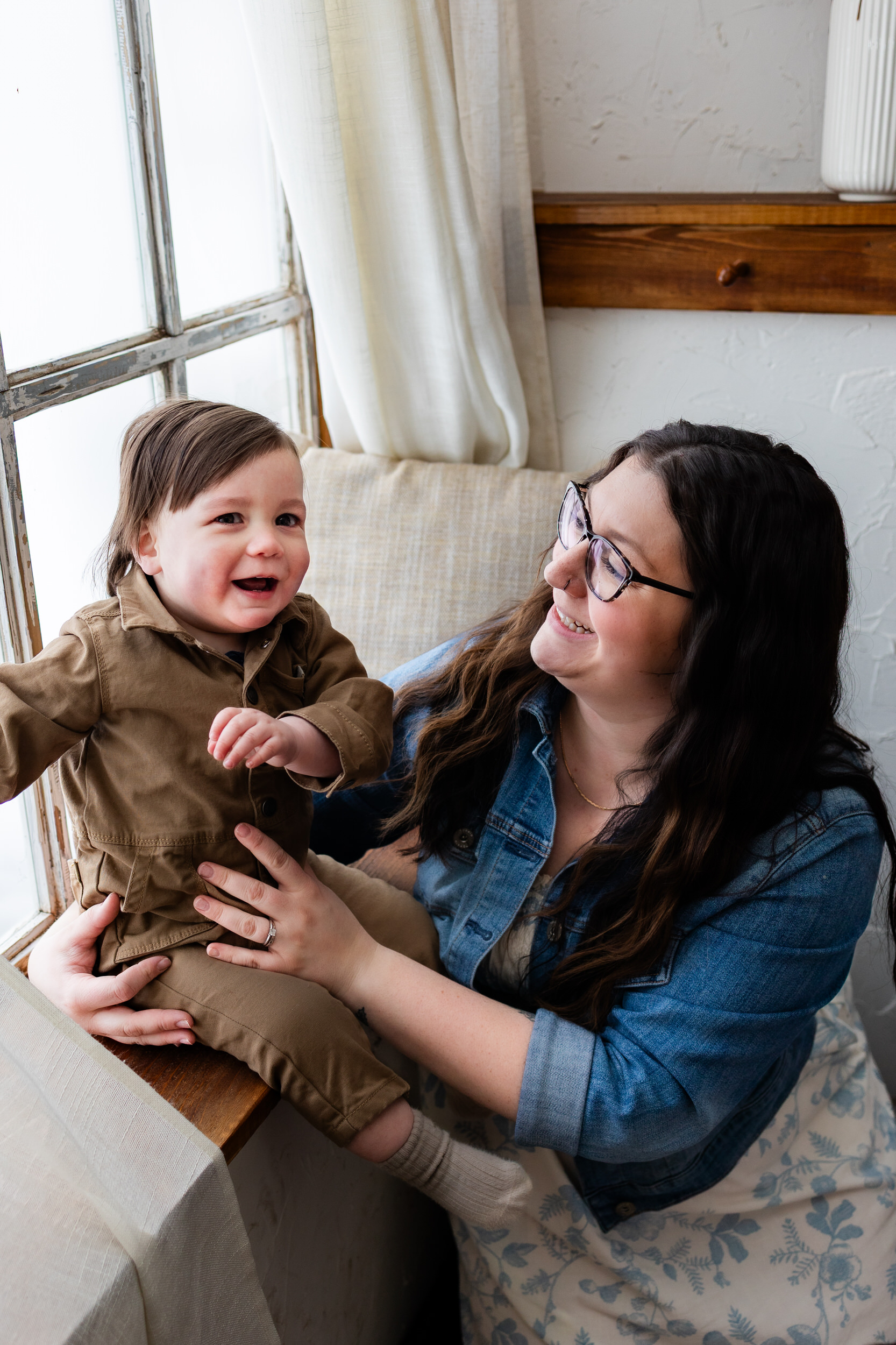 Little boy sits in a windowsill as mom holds onto him and smiles at him.