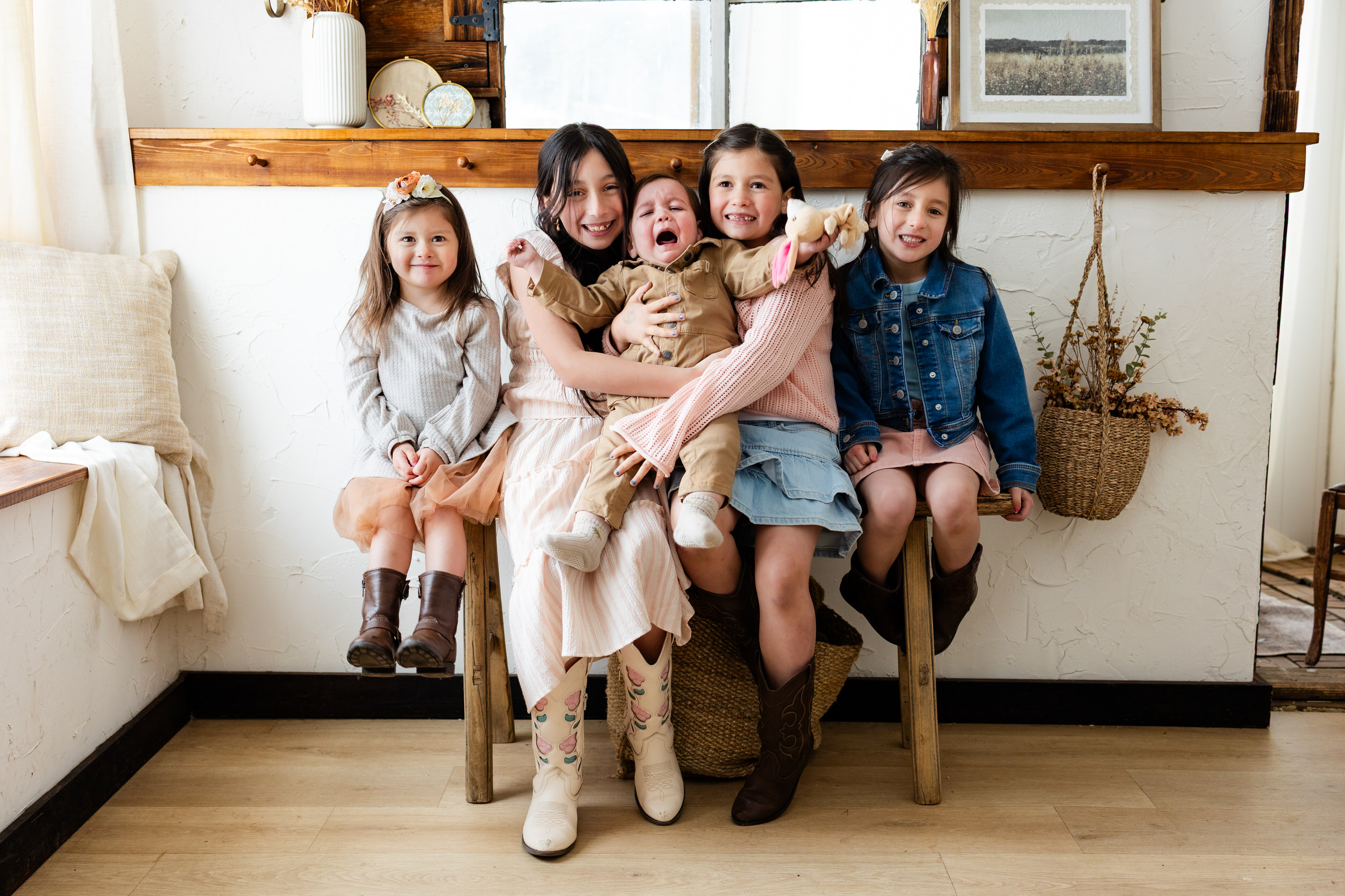 Four sisters sit on a bench in a cottage and hold their younger brother who is crying.