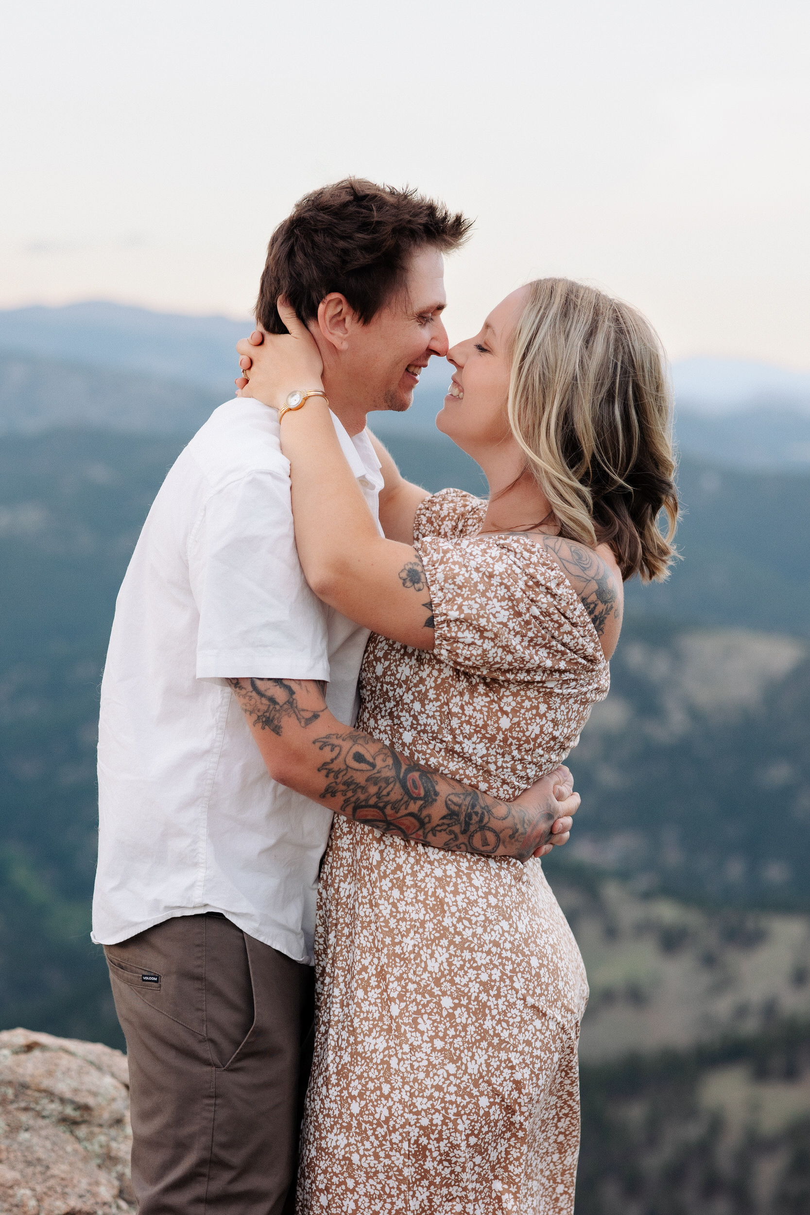Husband and wife laugh together and lean in for a kiss on a mountain top at Lost Gulch Overlook in Boulder, Colorado.