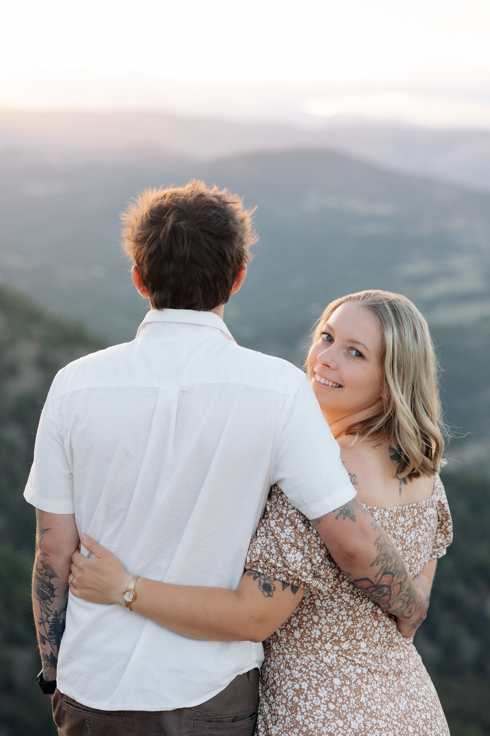 Husband and wife stand with their arms around each other's waist and wife looks back over her shoulder and smiles at the camera.