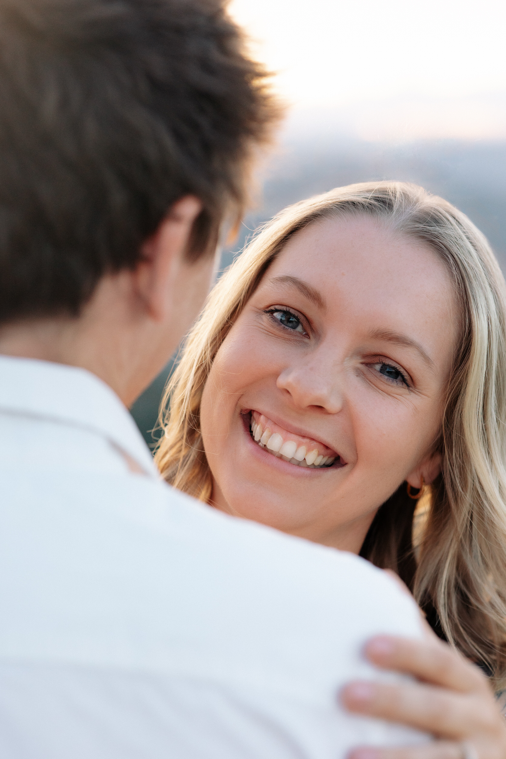 Wife peeks over her husband's shoulder and smiles at the camera.