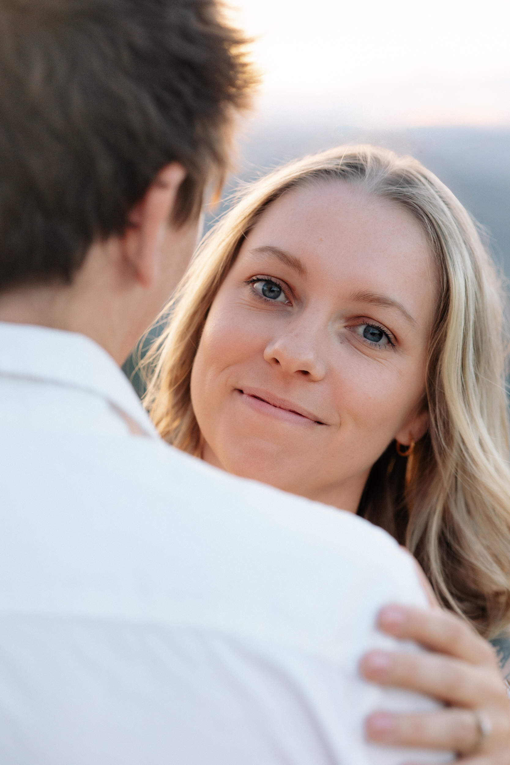 Wife peeks over her husband's shoulder at the camera.